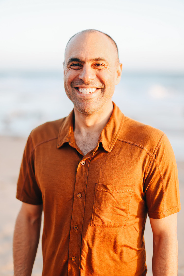 Ben Mati smiling on the beach in a rust-colored shirt, ocean and soft evening light behind
