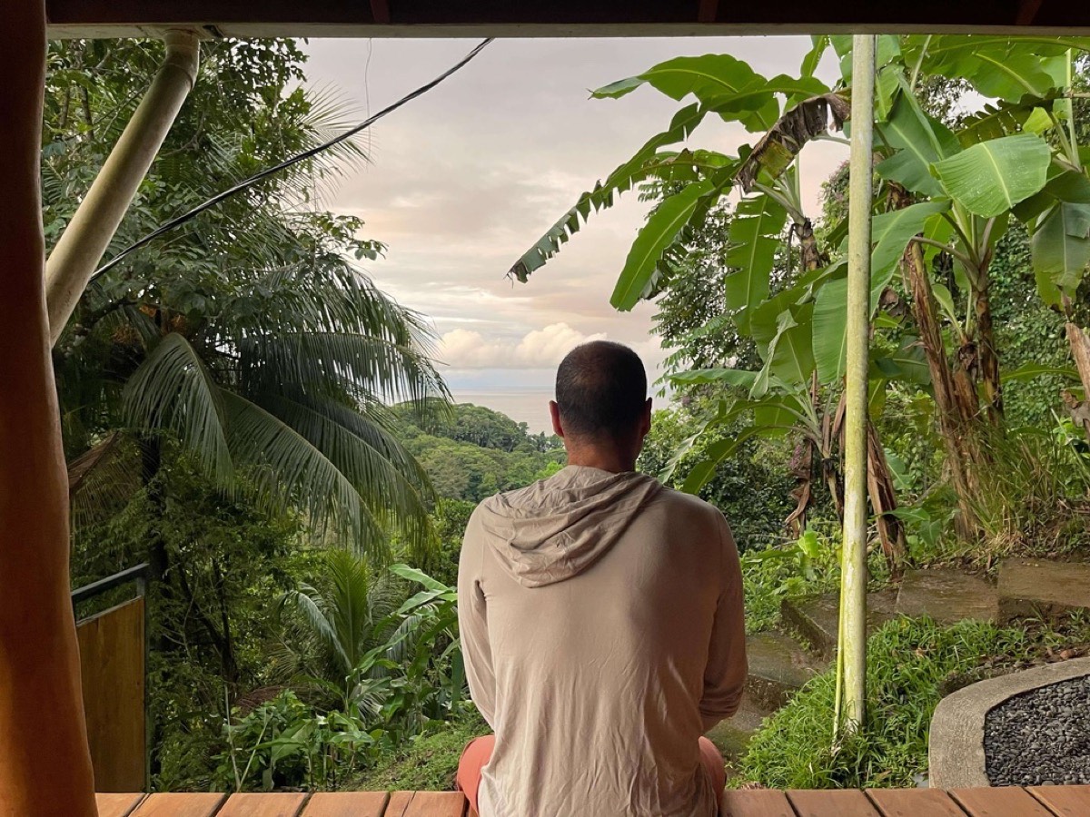 Ben seated on a wooden deck, seen from behind, looking out over tropical forest toward the ocean