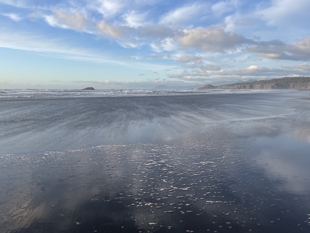 Wet sand at the water's edge reflecting the sky and clouds, with sea foam and a distant headland
